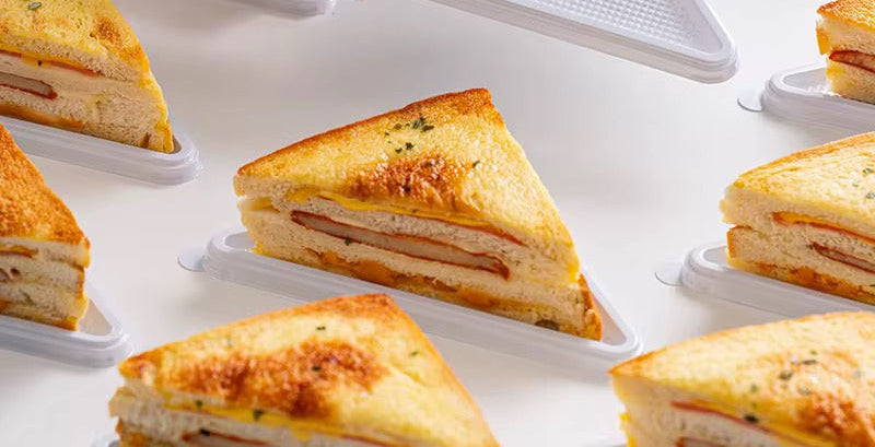 Person wearing gloves holding a sandwich above a tray with multiple sandwiches on a white surface. The business sells packaging for desserts.